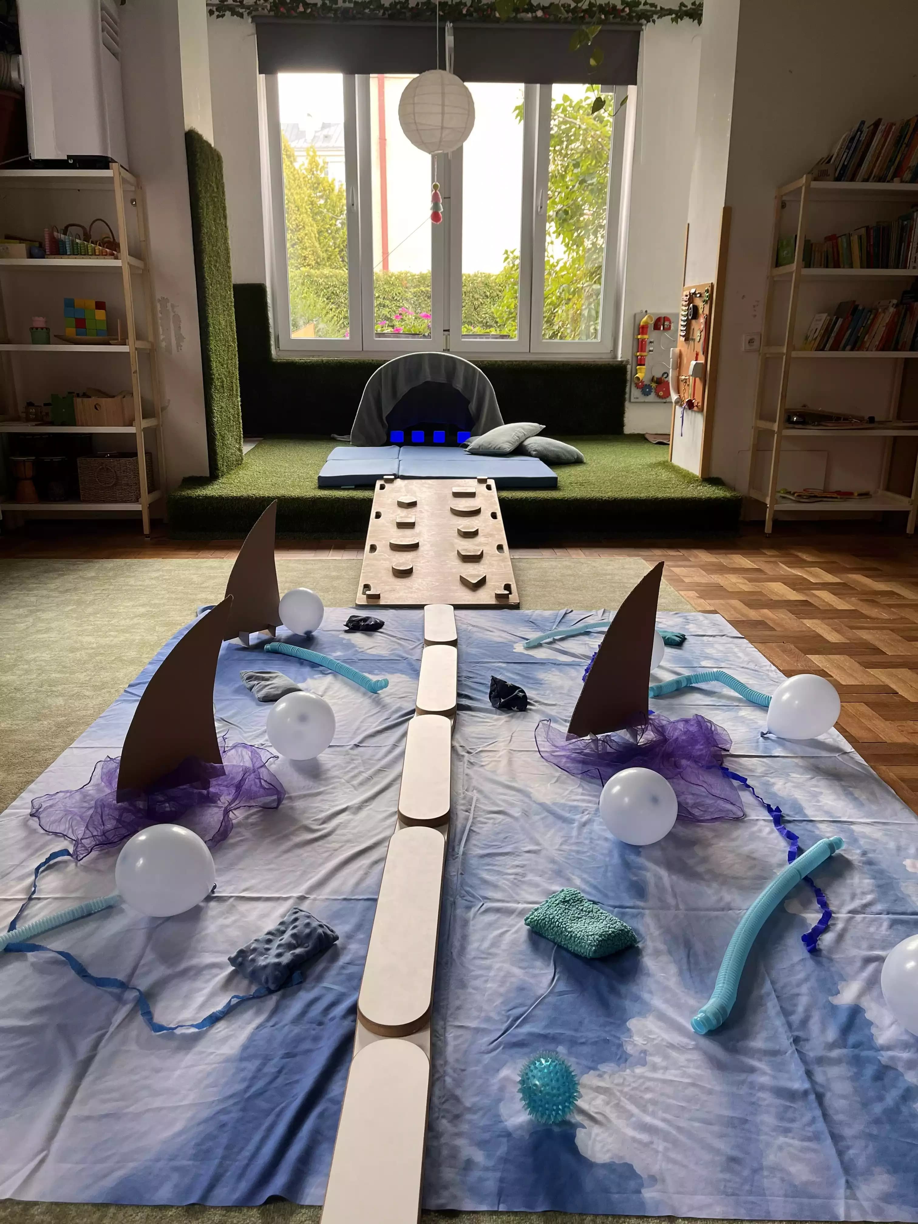 Sala żłobka z przygotowaną ścieżką sensoryczną. Na podłodze znajduje się niebieskie płótno z różnymi teksturami, balonami, oraz elementami, które dzieci mogą dotykać i eksplorować.